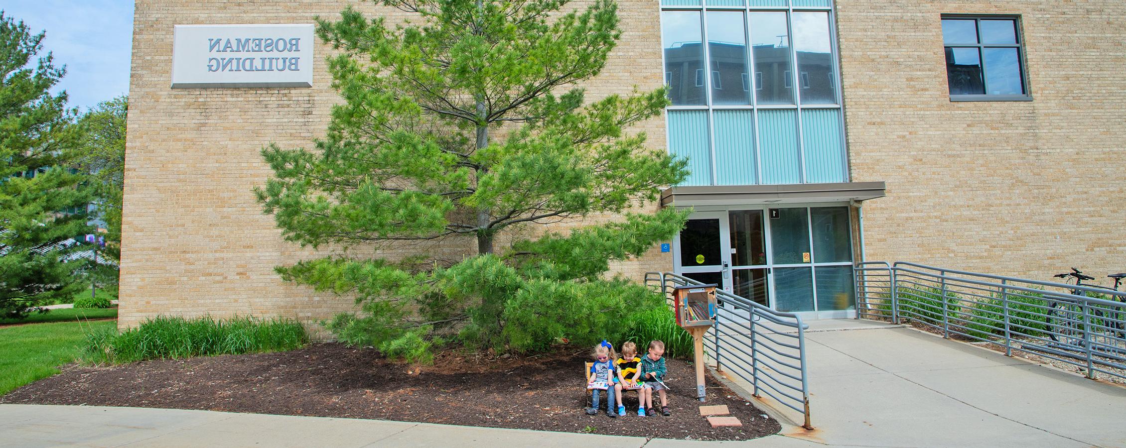 Image outside of Roseman hall with a green tree in front of it and three children sitting in front of the tree.