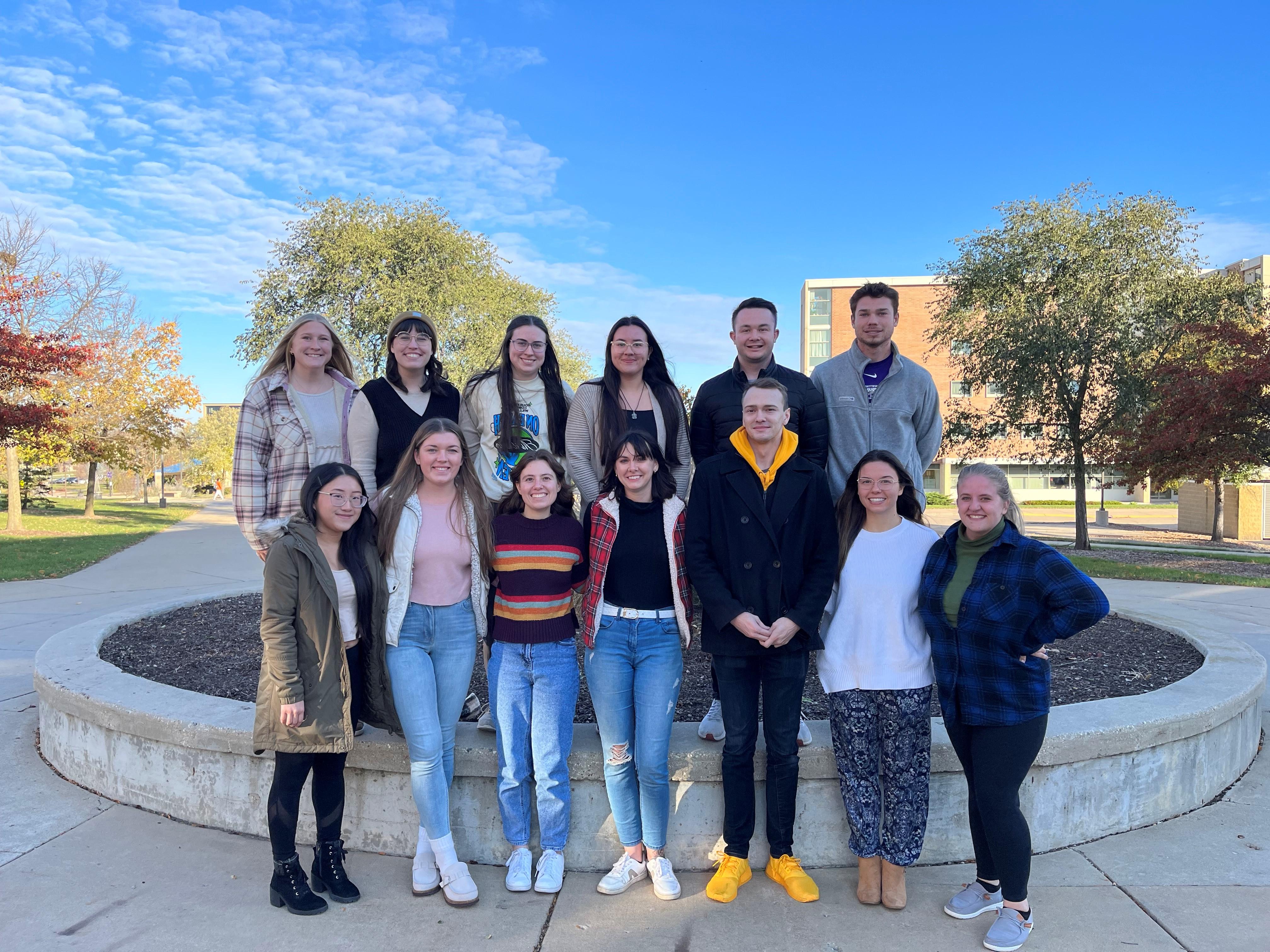 A group of people stand together in two rows outdoors on campus.