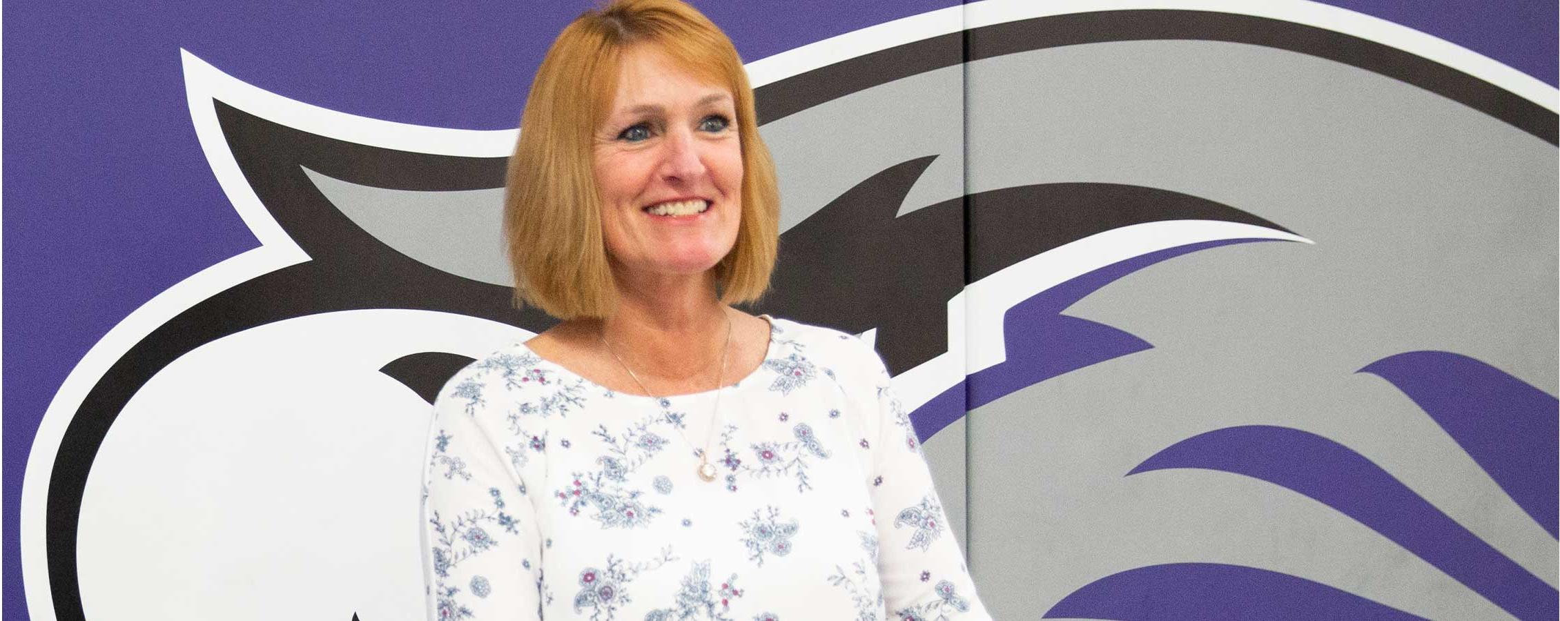 Kathleen Happel stands in front of a mat wall with a large Warhawk on it.